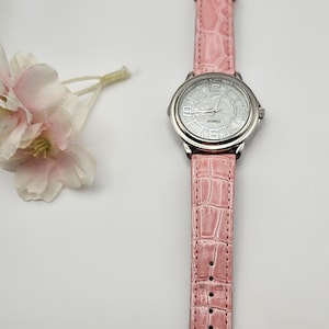 May include: A pink quartz watch with a silver-colored bezel and a matching pink faux-leather band. The watch face is white with black numerals and hands. The band has a crocodile texture. A small floral arrangement is in the background.