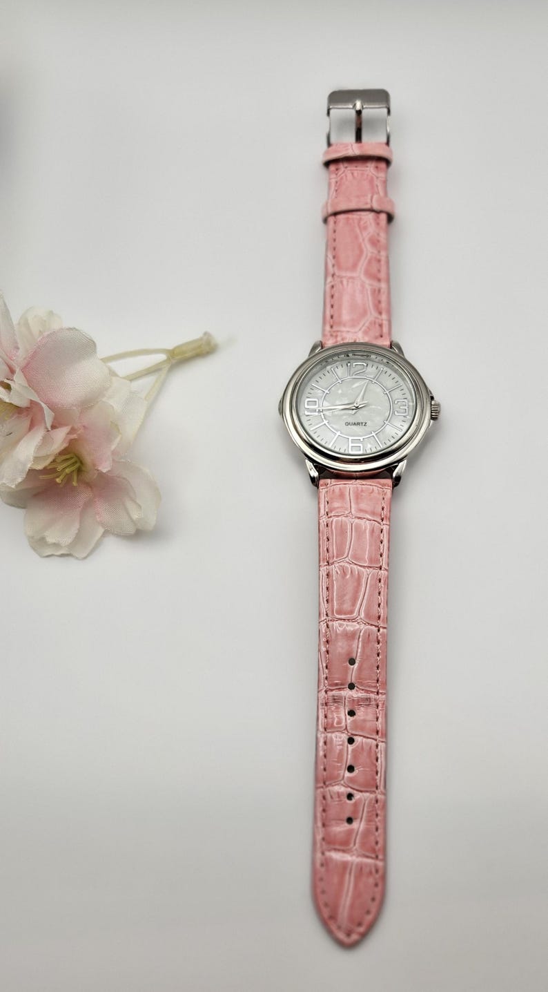 May include: A pink quartz watch with a silver-colored bezel and a matching pink faux-leather band. The watch face is white with black numerals and hands. The band has a crocodile texture. A small floral arrangement is in the background.