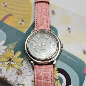 May include: A silver quartz watch with a light pink faux-leather band. The watch face is silver with black numerals and hands. The band has a crocodile skin texture. The watch is on a floral and bee-themed background.