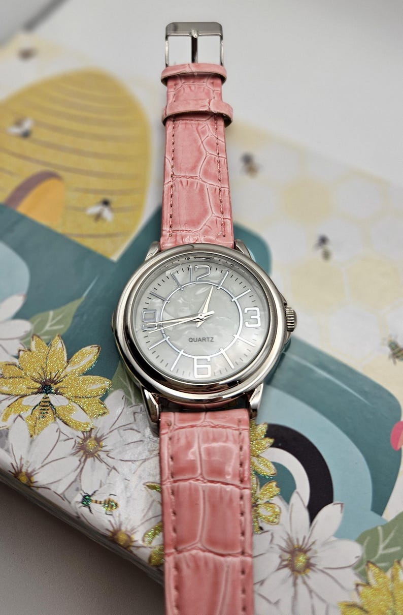 May include: A silver quartz watch with a light pink faux-leather band. The watch face is silver with black numerals and hands. The band has a crocodile skin texture. The watch is on a floral and bee-themed background.