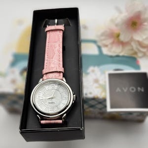May include: A silver-toned quartz watch with a light pink leather band, presented in a black box. The watch face is white with silver numerals and hands. The watch is from the brand Avon.