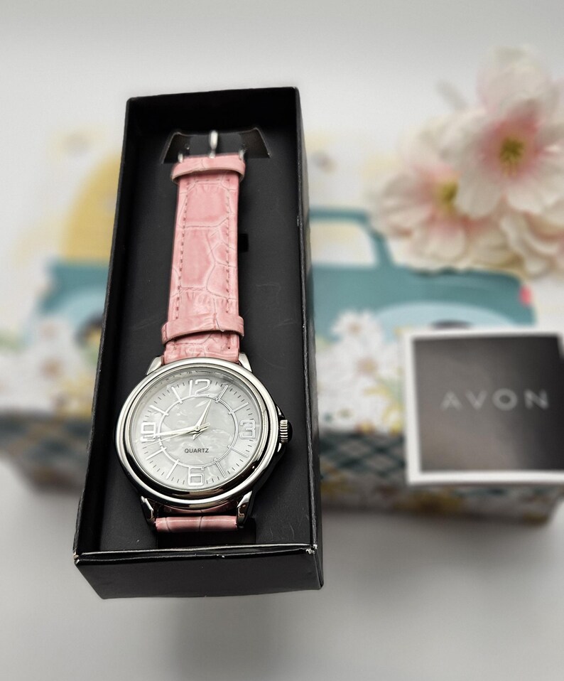 May include: A silver-toned quartz watch with a light pink leather band, presented in a black box. The watch face is white with silver numerals and hands. The watch is from the brand Avon.