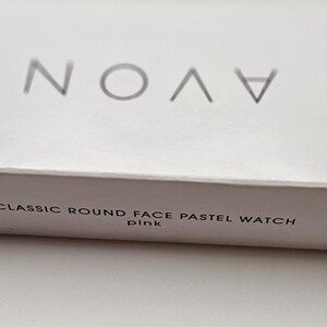 May include: A white box with the brand name "AVON" in silver lettering. The box also has the text "CLASSIC ROUND FACE PASTEL WATCH pink" printed in a smaller, gray font. The box is on a white surface.
