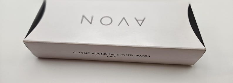 May include: A white box with the brand name "AVON" in silver lettering. The box also has the text "CLASSIC ROUND FACE PASTEL WATCH pink" printed in a smaller, gray font. The box is on a white surface.