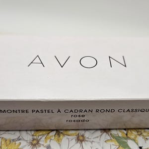 May include: A white box with the word "AVON" in black letters. The box has the text "MONTRE PASTEL &Agrave; CADRAN ROND CLASSIQUE rose rosado" printed on it. The box is sitting on a floral patterned surface.