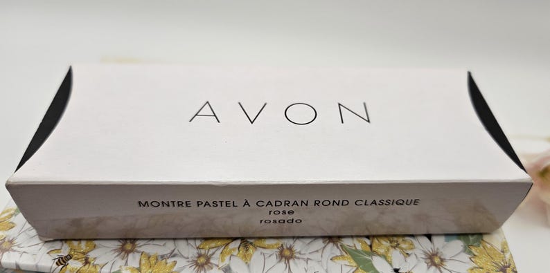 May include: A white box with the word "AVON" in black letters. The box has the text "MONTRE PASTEL &Agrave; CADRAN ROND CLASSIQUE rose rosado" printed on it. The box is sitting on a floral patterned surface.