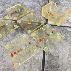 Könnte beinhalten: Set aus sechs quadratischen Untersetzern aus Harz und einem passenden Halter. Jeder Untersetzer ist transparent mit einem gelben Farbton und mit bunten Edelsteinen versehen. Der Halter hat ein ähnliches Design. Die Untersetzer haben kleine, runde, transparente Füße.