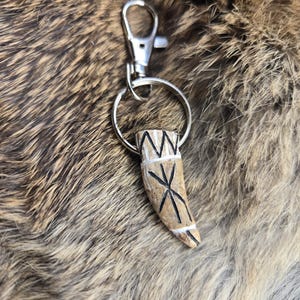May include: A keychain featuring a carved, bone-colored pendant with black and white markings. The pendant is shaped like a tooth and is attached to a silver-colored metal ring and clasp. The background is a textured, brown and gray fur.