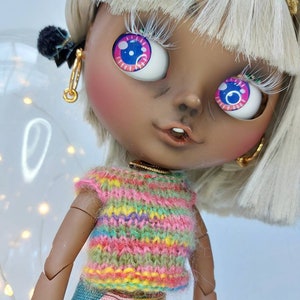 Könnte beinhalten: Eine Custom-Puppe mit gebräuntem Teint, weißem Haar und großen, ausdrucksstarken Augen mit rosa und blauen Iris. Die Puppe trägt ein bunt gestreiftes Strickoberteil und einen gemusterten Rock. Goldene Creolen und eine goldene Krone.