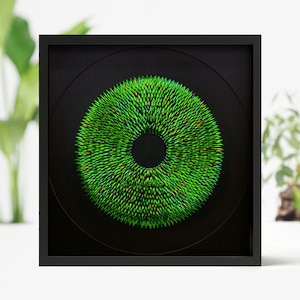May include: A framed circular artwork with a black background and a green and red ring of small, pointed objects. The ring is centered around a black circle.