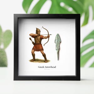 May include: A framed print depicting a Greek archer with a bow and arrow, and a bronze arrowhead. The archer is wearing armour. The text "Greek Arrowhead" and dates 323-31 BC are below the image. The frame is black.