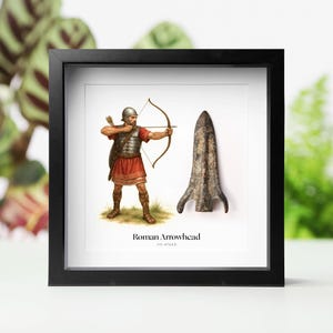 May include: Framed print featuring a Roman soldier drawing a bow and arrow, alongside a detailed illustration of a Roman arrowhead. The text "Roman Arrowhead" and the date range 110-476 AD are displayed below the images. The frame is black.