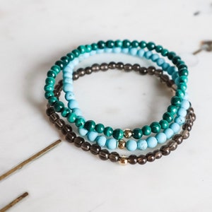 May include: Three beaded bracelets stacked on a white surface. The top bracelet is green, the middle bracelet is light blue, and the bottom bracelet is brown.
