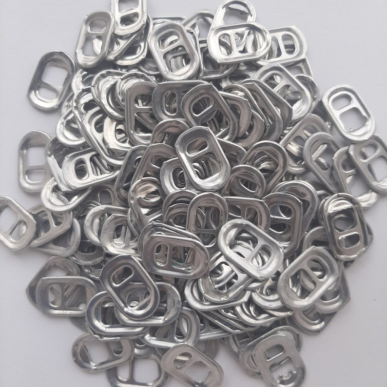May include: A large pile of silver pull tabs from aluminum beverage cans. The tabs are all the same size and shape, and they are arranged in a random pattern.