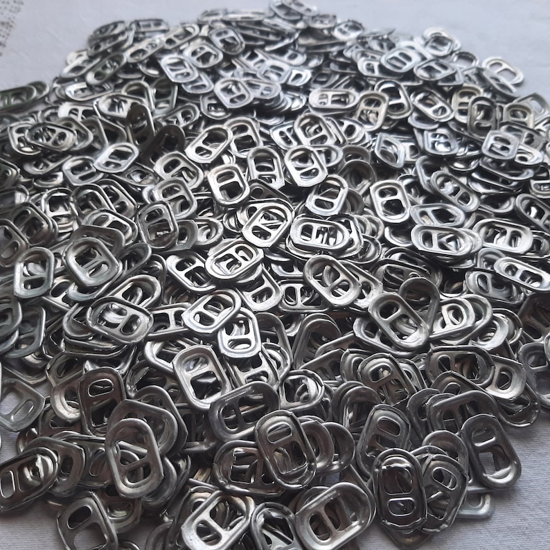 May include: A large pile of silver pull tabs from aluminum beverage cans. The tabs are arranged in a random pattern, creating a textured surface.