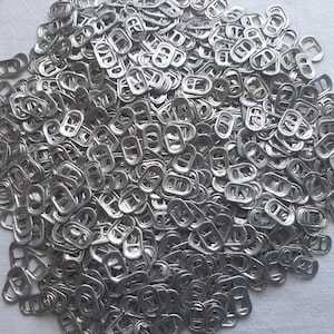 May include: A large pile of silver pull tabs from aluminum beverage cans. The tabs are all the same size and shape, and they are arranged in a random pattern.