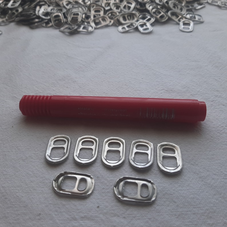 May include: A close-up of seven silver pull tabs from aluminum cans. The pull tabs are arranged in two rows on a white surface. A red marker is visible above the pull tabs.