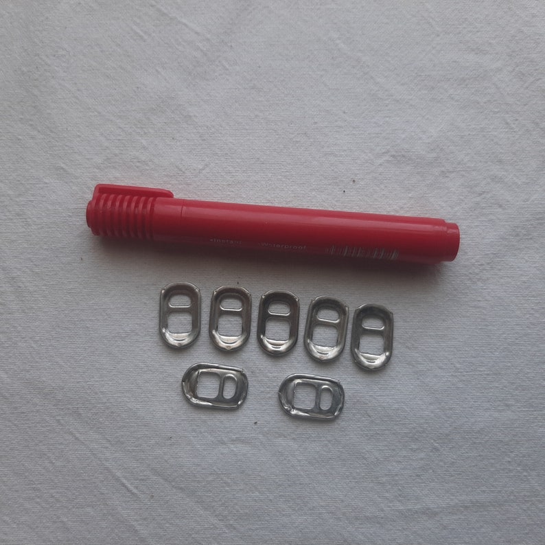 May include: A red marker pen and seven silver pull tabs from aluminum beverage cans. The pull tabs are arranged in two rows on a white surface.