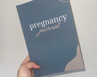 May include: A blue pregnancy journal with the words "pregnancy journal" in white and a light brown accent design. The journal is held in a hand against a white background.