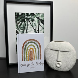 May include: A white journal with a rainbow design and the text "Bump to Baby Journal". The journal is sitting on a black surface next to a white ceramic vase with a face design.