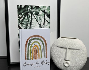 May include: A white journal with a rainbow design and the text "Bump to Baby Journal". The journal is sitting on a black surface next to a white ceramic vase with a face design.