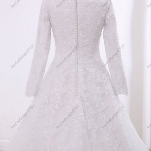 May include: A white wedding dress with long sleeves and a lace overlay. The dress has a fitted bodice and a flowing skirt. The back of the dress features a row of buttons.