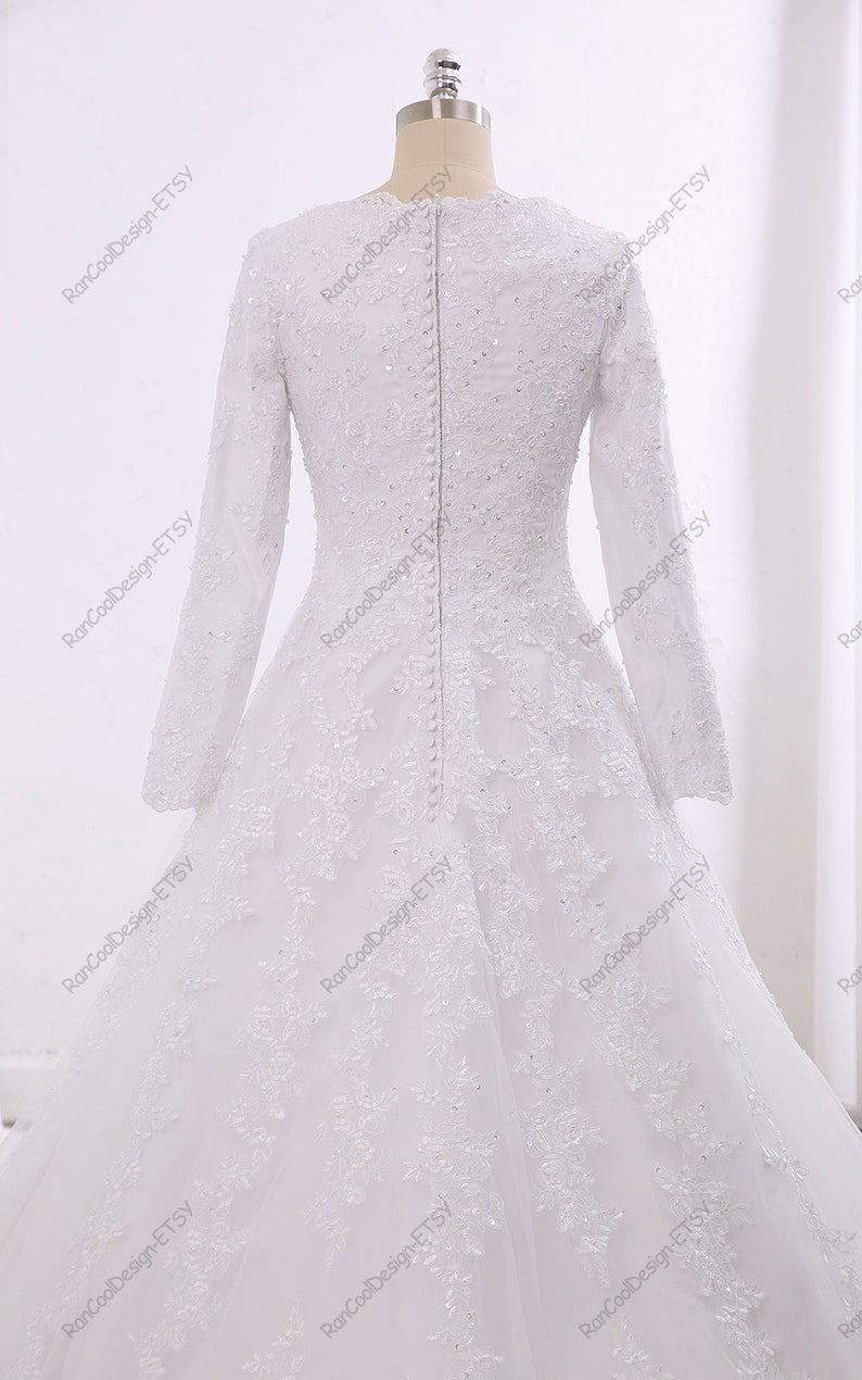 May include: A white wedding dress with long sleeves and a lace overlay. The dress has a fitted bodice and a flowing skirt. The back of the dress features a row of buttons.