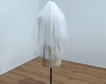 May include: A white, two-tiered bridal veil made of sheer fabric. The veil is displayed on a mannequin, showcasing its layered design and delicate texture. The veil is a classic accessory for weddings.