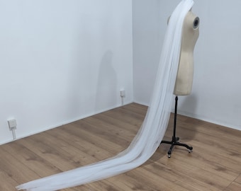 May include: A long, flowing white bridal veil displayed on a mannequin. The veil is made of a sheer, lightweight fabric and drapes down to the floor. The mannequin is a neutral beige color, and the background is a plain white wall and wooden floor.