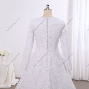 May include: A white wedding dress with long sleeves and a lace bodice. The dress has a fitted bodice and a flowing skirt. The dress is decorated with delicate beading and lace.