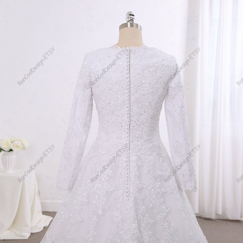 May include: A white wedding dress with long sleeves and a lace bodice. The dress has a fitted bodice and a flowing skirt. The dress is decorated with delicate beading and lace.