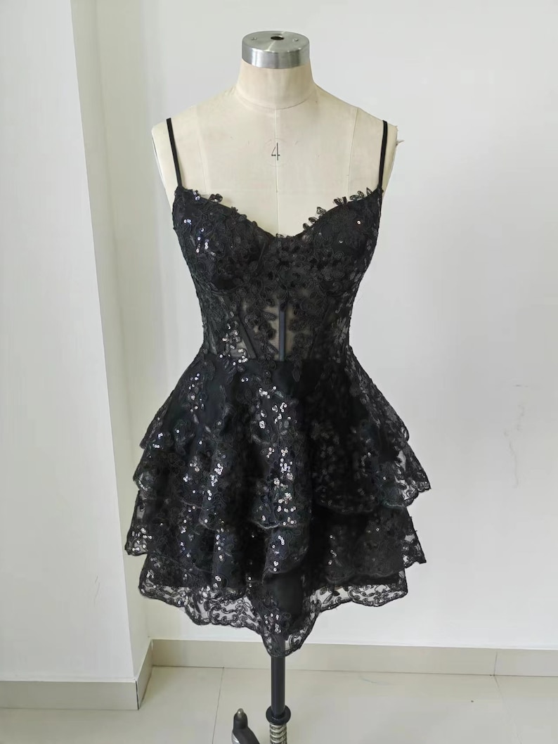 May include: A black short dress with a layered skirt and sequins. The dress has a sweetheart neckline and spaghetti straps.
