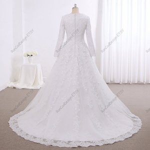 May include: A white wedding dress with long sleeves and a lace overlay. The dress has a fitted bodice and a full skirt. The back of the dress features a row of buttons.