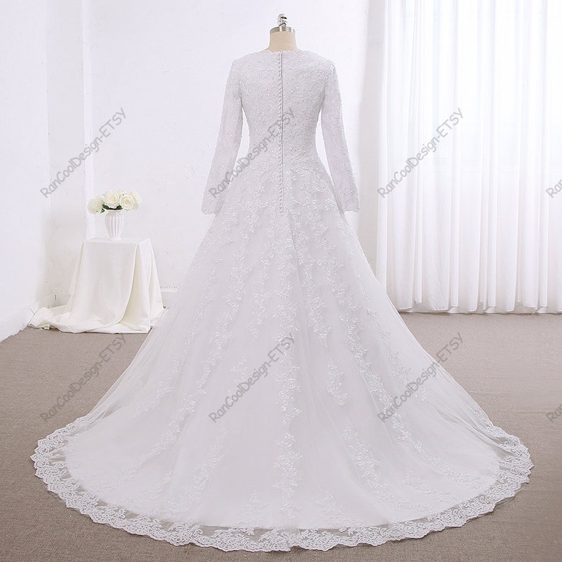 May include: A white wedding dress with long sleeves and a lace overlay. The dress has a fitted bodice and a full skirt. The back of the dress features a row of buttons.