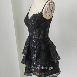 May include: A black short dress with a lace and sequin design. The dress has a sweetheart neckline and a layered skirt. The dress is on a mannequin.