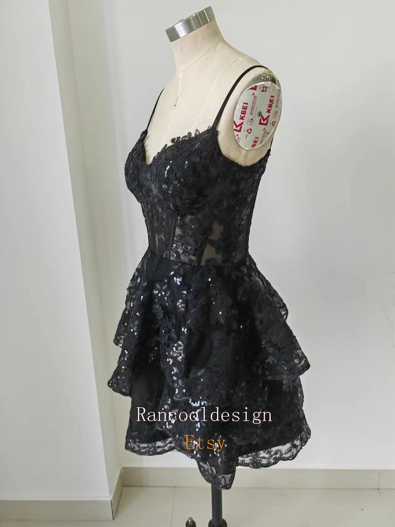 May include: A black short dress with a lace and sequin design. The dress has a sweetheart neckline and a layered skirt. The dress is on a mannequin.