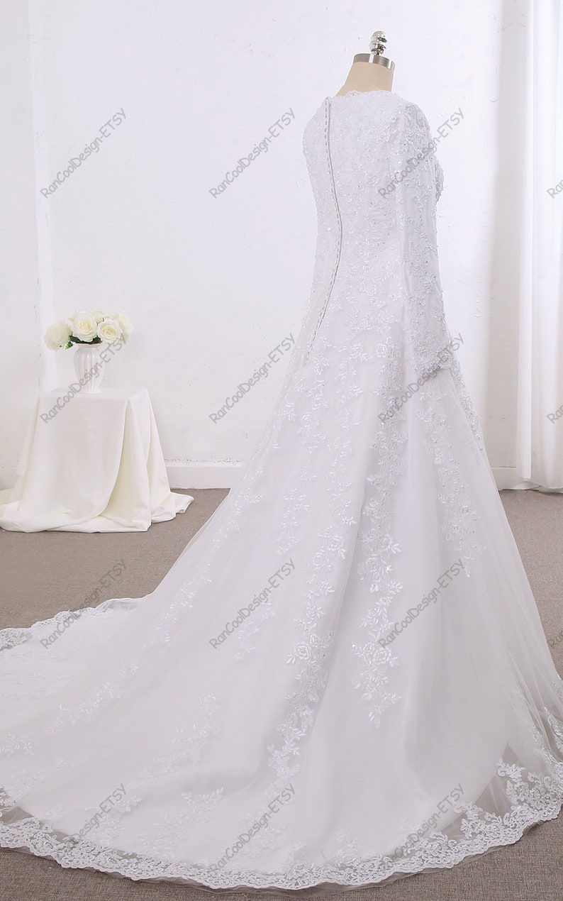 May include: A white wedding dress with long sleeves and a lace overlay. The dress has a fitted bodice and a flowing skirt with a train.