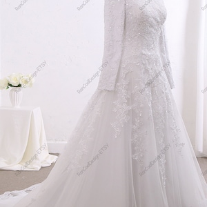 May include: A white wedding dress with long sleeves and a lace overlay. The dress has a fitted bodice and a flowing skirt. The skirt has a layer of tulle underneath the lace.