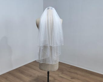 May include: A white, two-tiered bridal veil. The veil is made of a sheer, lightweight fabric and is displayed on a mannequin. The veil has a simple, elegant design, suitable for weddings and bridal events.