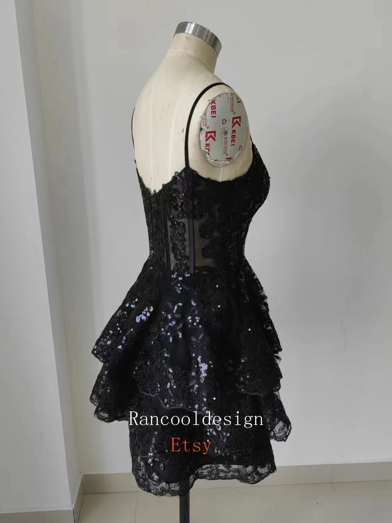 May include: A black short dress with a lace bodice and a layered skirt. The skirt has a ruffled design and is adorned with sequins. The dress is on a mannequin and the text "Rancooldesign Etsy" is visible at the bottom.