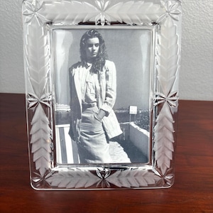 May include: A rectangular clear glass picture frame featuring a frosted leaf pattern etched along the border. The frame contains a black and white photograph of a person wearing a blazer and skirt, standing outdoors near a low wall and railing.