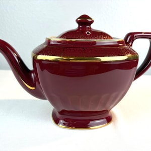 May include: A deep burgundy ceramic teapot featuring a gold-trimmed rim and a matching lid. The upper portion of the teapot body and the lid are decorated with a fine, gold-colored crackle pattern. It has a curved spout and a sturdy handle.