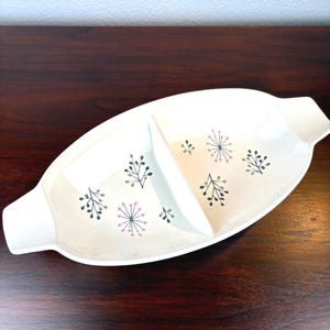 May include: A white oval serving dish with a divided interior. The dish is decorated with a pattern of black, gray, and pink floral designs. It has handles on each end and is set on a dark wood surface.