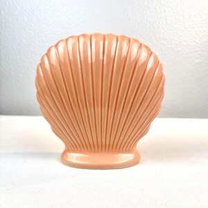May include: A peach-colored ceramic vase shaped like a scallop shell. The object features a glossy finish with vertical ridges radiating from the base to the scalloped top edge. It has a wide, rounded body and a stable, flat circular base.