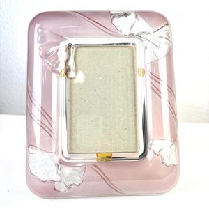 May include: A rectangular picture frame made of translucent pink glass. The frame features clear, embossed ginkgo leaf motifs at the corners and curved, raised glass lines that sweep across the surface. A central rectangular opening holds a beige insert.