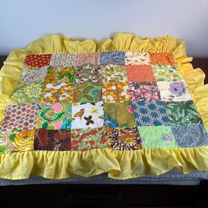 May include: A patchwork quilt with a yellow ruffled border. The quilt is made of various fabric squares with floral, geometric, and abstract patterns in shades of red, green, blue, and orange. The quilt is approximately 30 inches wide and 30 inches long.