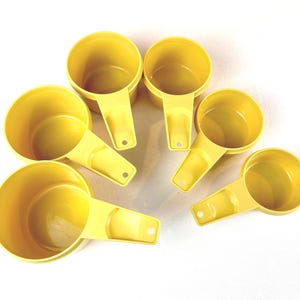 May include: A set of five bright yellow plastic measuring cups of varying sizes, each featuring a round bowl and a flat handle with a small circular hole at the end for hanging. The cups are arranged in a semi-circular pattern on a white surface.