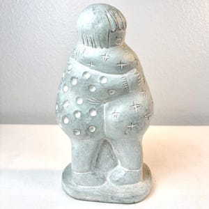 May include: A light gray stone sculpture depicting two figures in a close embrace. The figures are carved with textured details, including circular polka dots on one side and small cross-shaped patterns on the other, standing on a base.