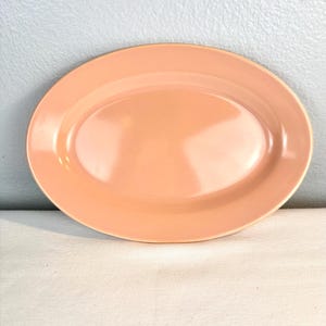 May include: A peach-colored oval serving platter. The ceramic dish has a glossy finish and a slightly raised rim. The platter is empty and sits on a white surface against a light blue wall.