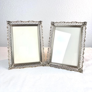 May include: Two rectangular picture frames with a metallic silver finish. Each frame features a decorative lattice or filigree border pattern surrounding a central glass pane. The frames are standing upright, showcasing a clear, reflective surface.